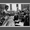 Black-and-white magazine photo of a woman in a patterned dress among seated people.