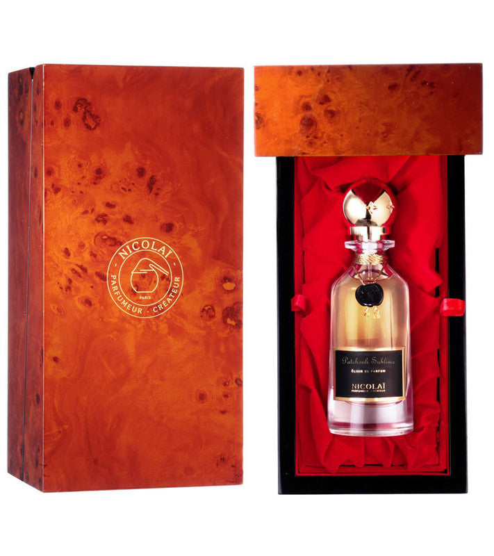 A glass perfume bottle with a gold cap labeled "Nicolaï" is displayed in an open wooden box with a red interior lining. The box, adorned with the Nicolaï logo, holds the exquisite Patchouli Sublime Elixir, known for its rich patchouli fragrance notes.
