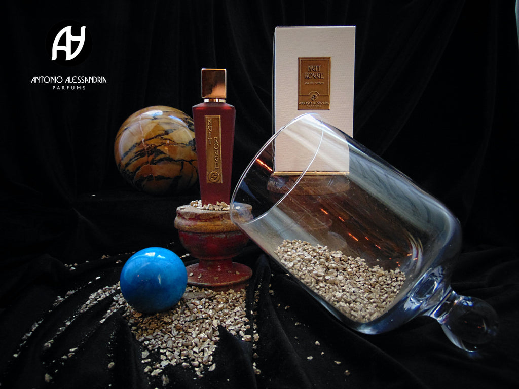 A display of Antonio Alessandria Nuit Rouge includes a red perfume bottle, white box, glass jar spilling granules, and decorative spherical objects on a black fabric backdrop. The collection hints at the rich leather scent and spicy top notes that define these exquisite fragrances.