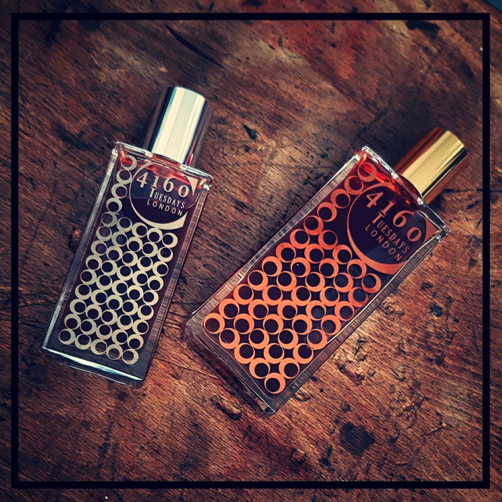 Two perfume bottles of Over the Chocolate Shop from 4160Tuesdays, featuring a circular pattern on the front, placed on a wooden surface. Their captivating fragrance is complemented by one silver cap and one gold cap, making them as delightful to the eyes as an aroma-rich visit to a chocolate shop.