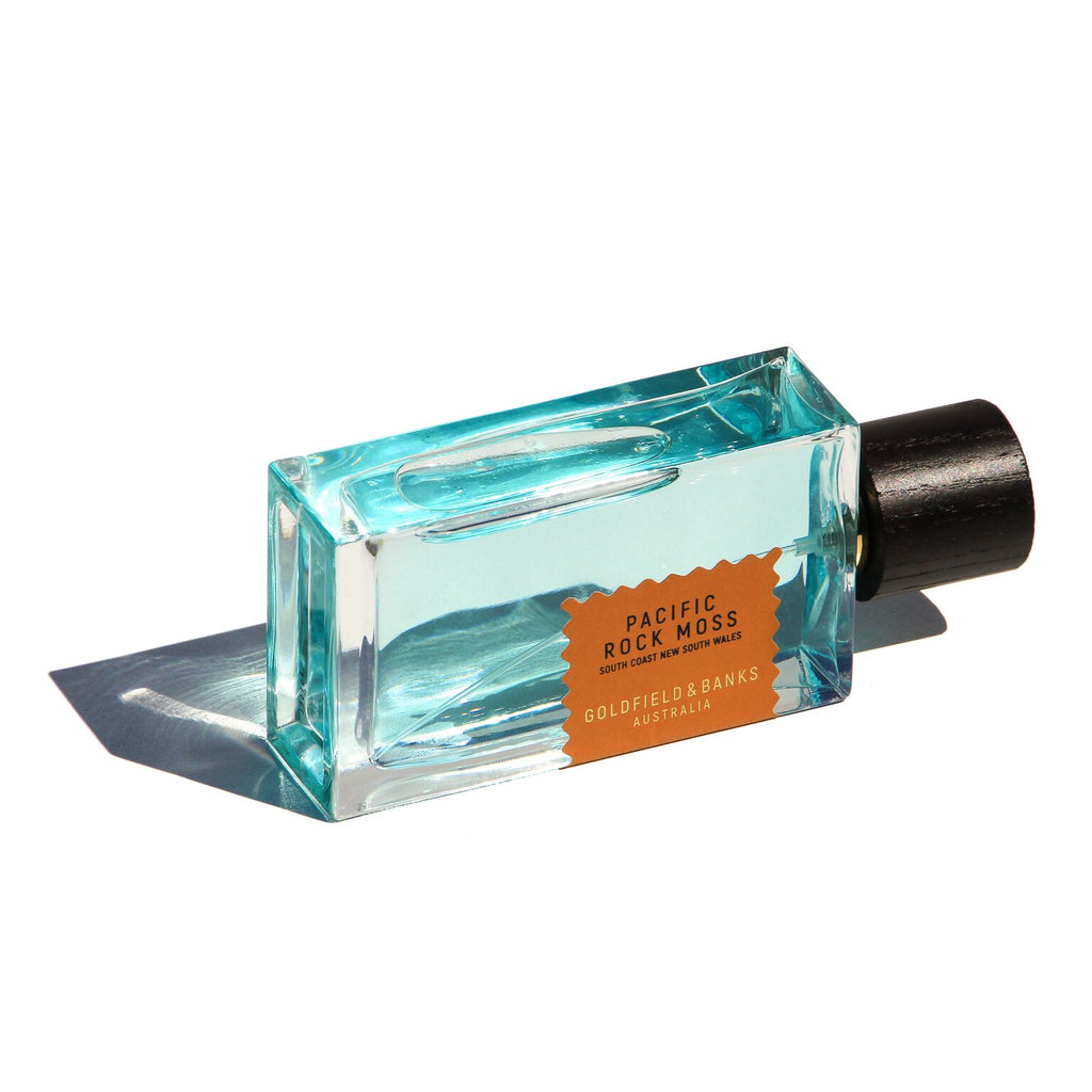 A rectangular perfume bottle labeled "Pacific Rock Moss" by Goldfield & Banks, filled with turquoise liquid and featuring a black cap, lies on its side against a white background, embodying the essence of a fresh summer fragrance perfect for embracing beach life.