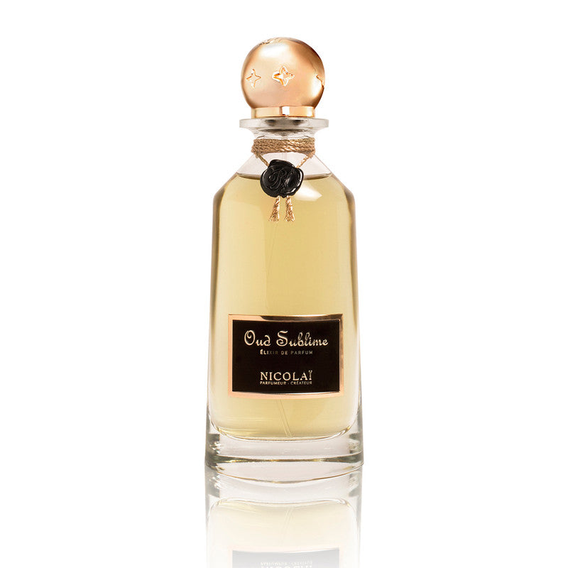 A clear glass bottle of Oud Sublime Elixir by Nicolaï, featuring a gold cap and black label, embodies the essence of Cambodian oud.