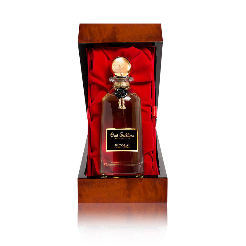 A glass bottle of Oud Sublime Elixir by Nicolaï, infused with hints of Cambodian oud, placed in an open wooden box lined with red fabric.