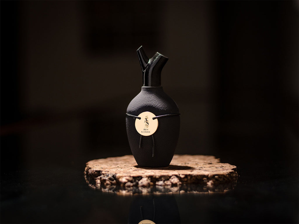 The Plum In Cognac by Scents of Wood, a small black vase-shaped bottle with a gold emblem, is displayed on a round rustic wooden surface against a dark background, exuding aromas of dark rum and vanilla.