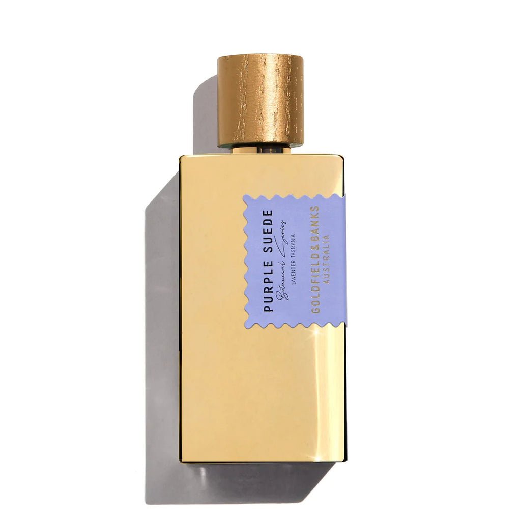 The Golden rectangular perfume bottle from Goldfield & Banks features a textured gold cap and a light purple label that reads "Purple Suede" and "Goldfield & Banks Australia." Capturing the essence of Lavender Tasmania, this exquisite Purple Suede perfume embodies the charm of luxurious leather. The bottle casts a shadow to the left.
