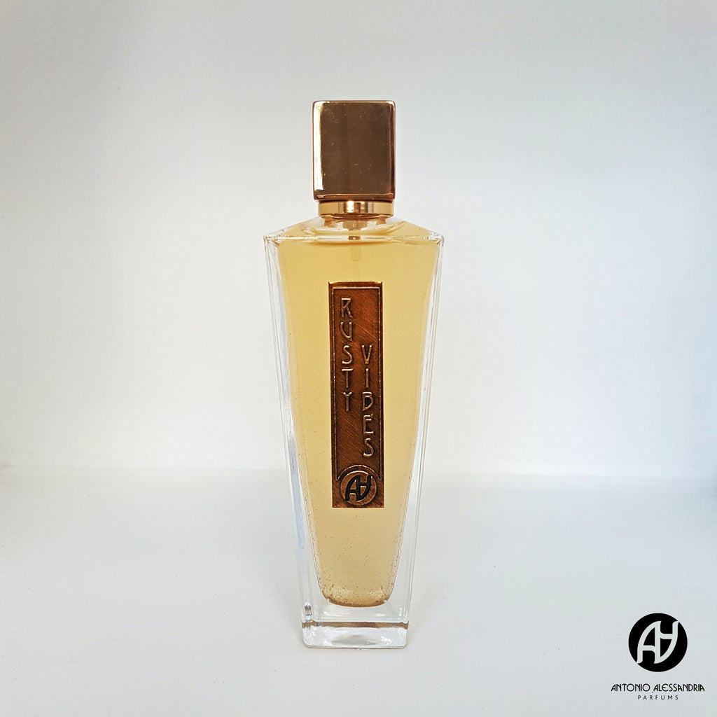 A clear glass bottle with a gold cap, labeled "Rusty Vibes," contains a yellow liquid with musky notes. The logo at the base reads "AY, Antonio Alessandria.