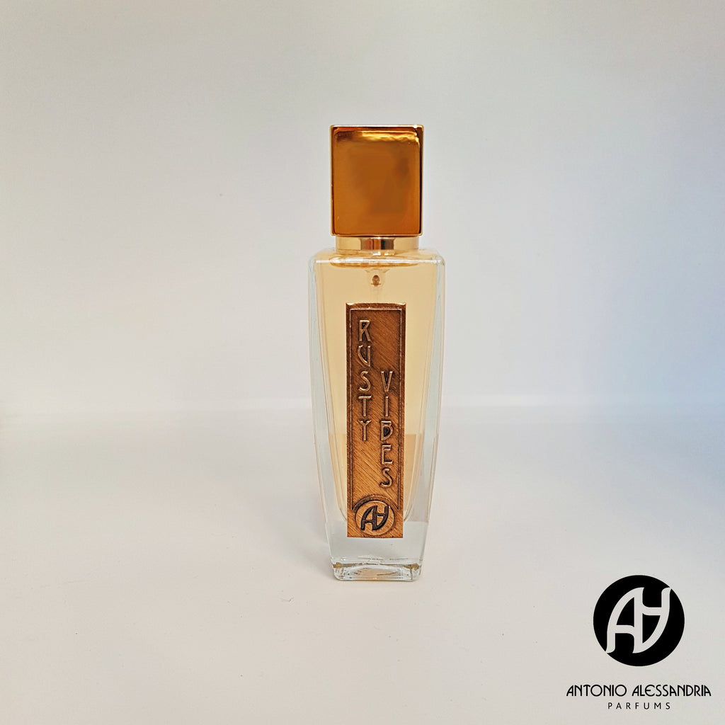 A clear bottle of floral perfume labeled "Rusty Vibes" with a gold cap showcases the refined craftsmanship of Antonio Alessandria. The brand name is subtly positioned at the bottom right, adding an elegant touch to this exquisite fragrance.
