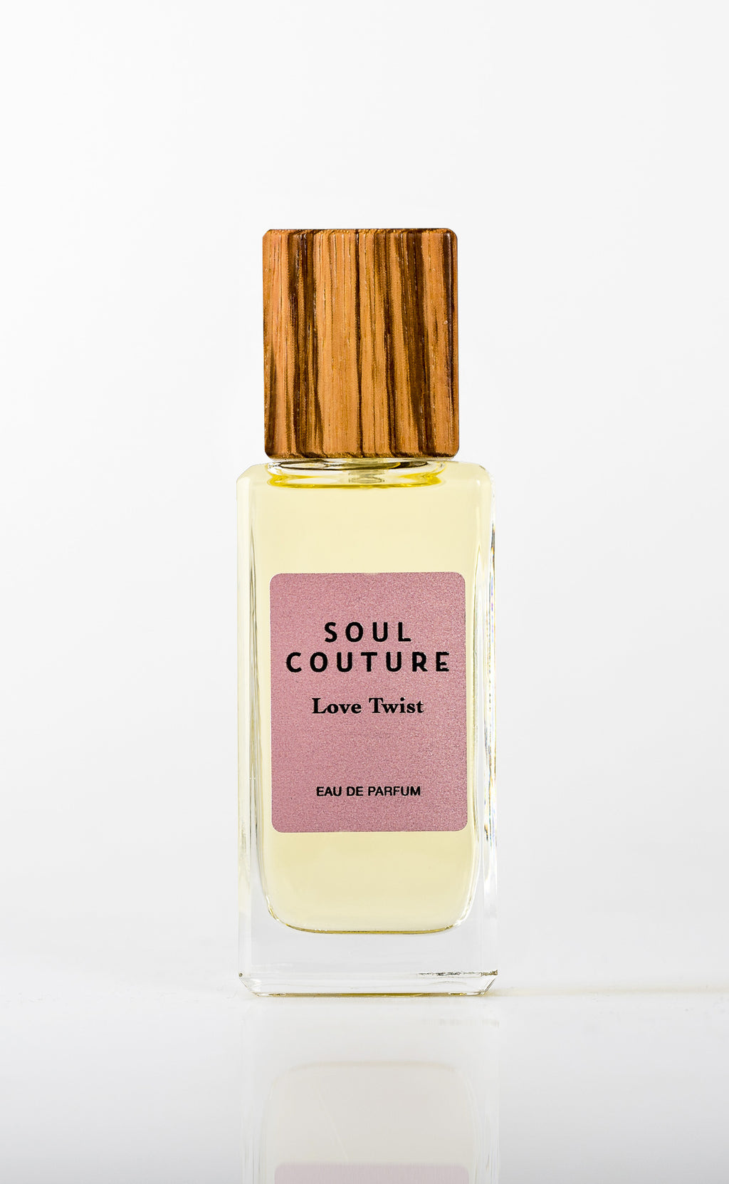 A bottle of Soul Couture Love Twist, enhanced with Ylang Ylang and featuring a wooden cap and pink label, rests on a white background.