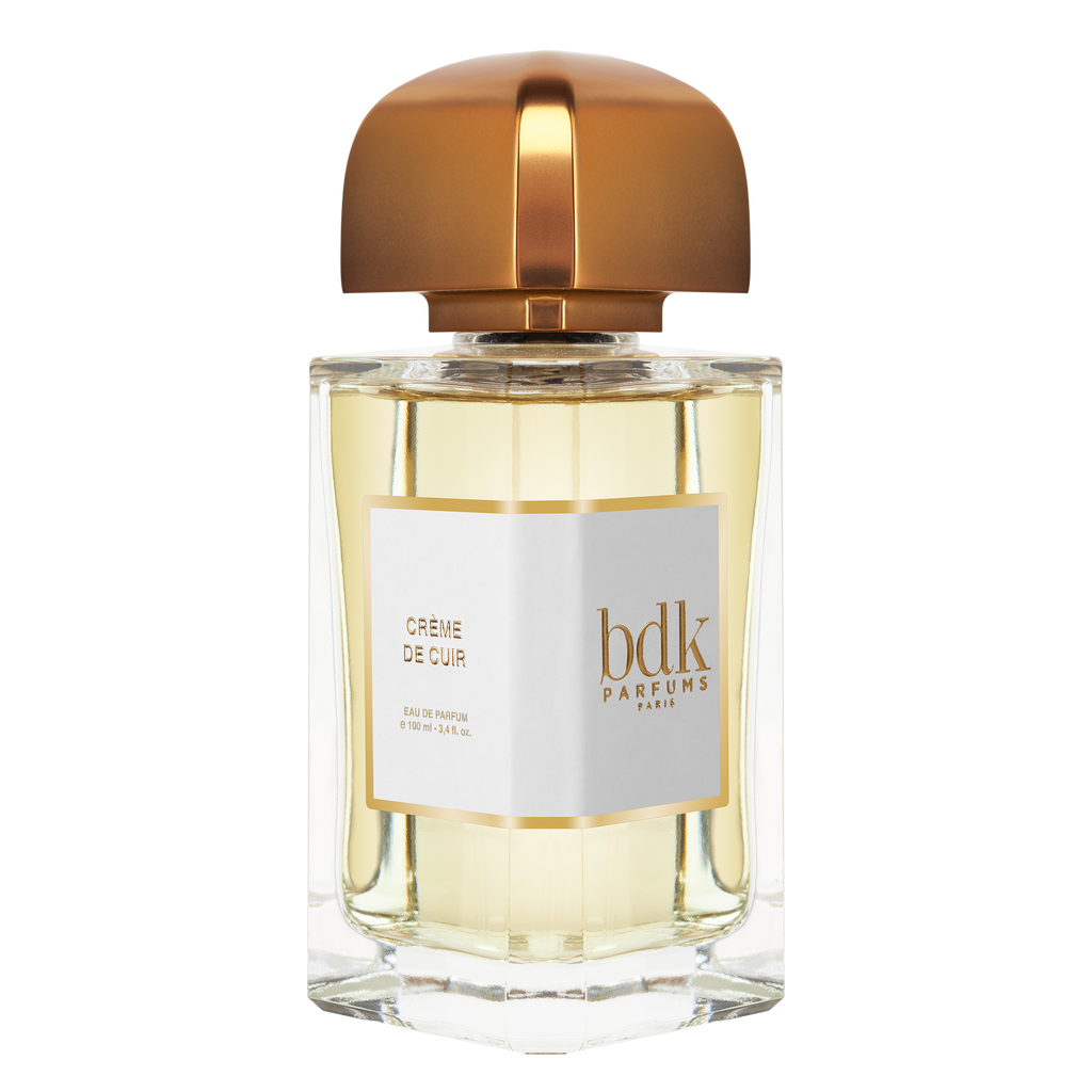 A bottle of Crème de Cuir by BDK Parfums, exuding contemporary sensuality with its golden cap and label on a clear glass container filled with light yellow liquid, reminiscent of an Indian summer.