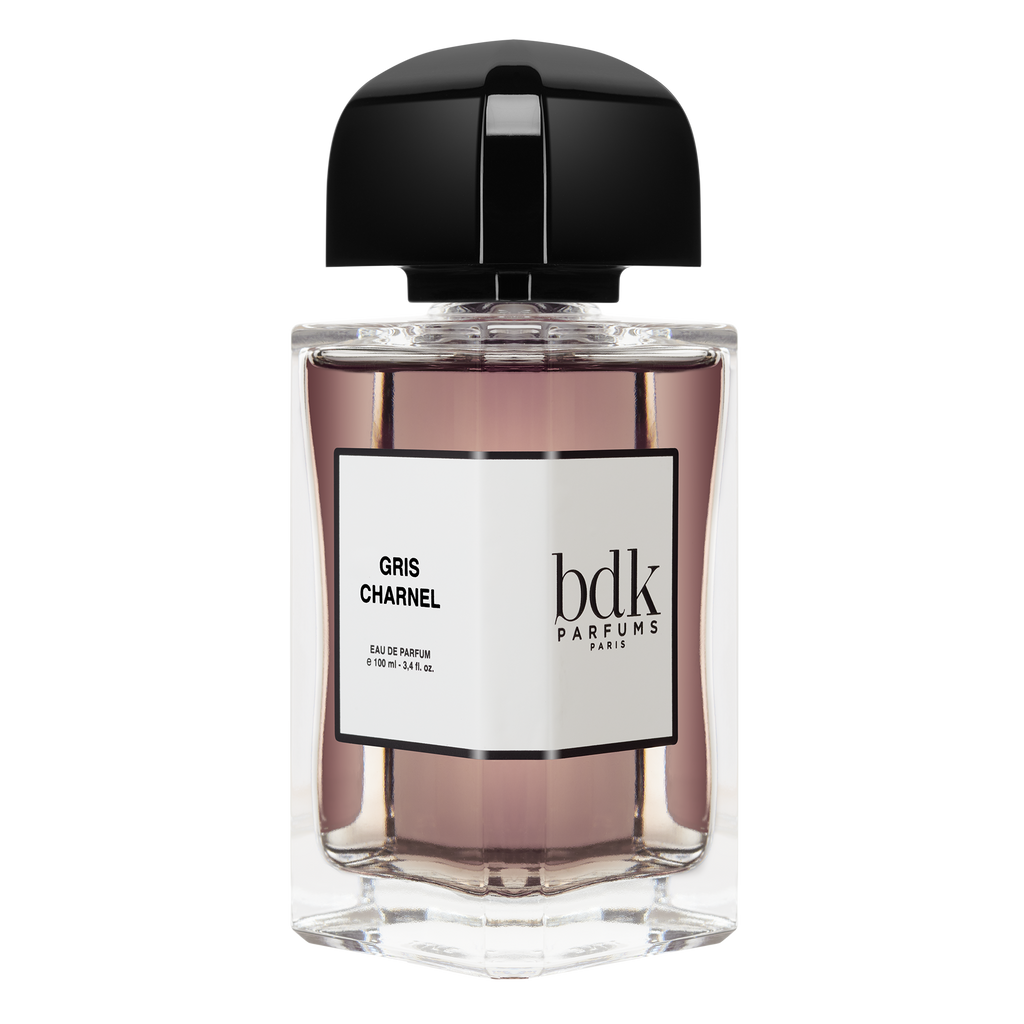 A clear glass bottle of Gris Charnel by BDK Parfums features a black cap and a white label displaying the perfume's name and brand. The liquid inside is a light pinkish-brown color, evoking the essence of sandalwood from India.