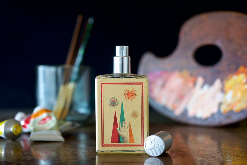 A bottle of Slow Explosions by Imaginary Authors with a colorful, abstract label is placed on a reflective surface. Nearby, paintbrushes in a glass, a painter's palette with mixed colors, and paint tubes create an artistic scene that hints at the rich notes of saffron and rose absolute within the scent.