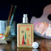 A bottle of Slow Explosions by Imaginary Authors with a colorful, abstract label is placed on a reflective surface. Nearby, paintbrushes in a glass, a painter's palette with mixed colors, and paint tubes create an artistic scene that hints at the rich notes of saffron and rose absolute within the scent.