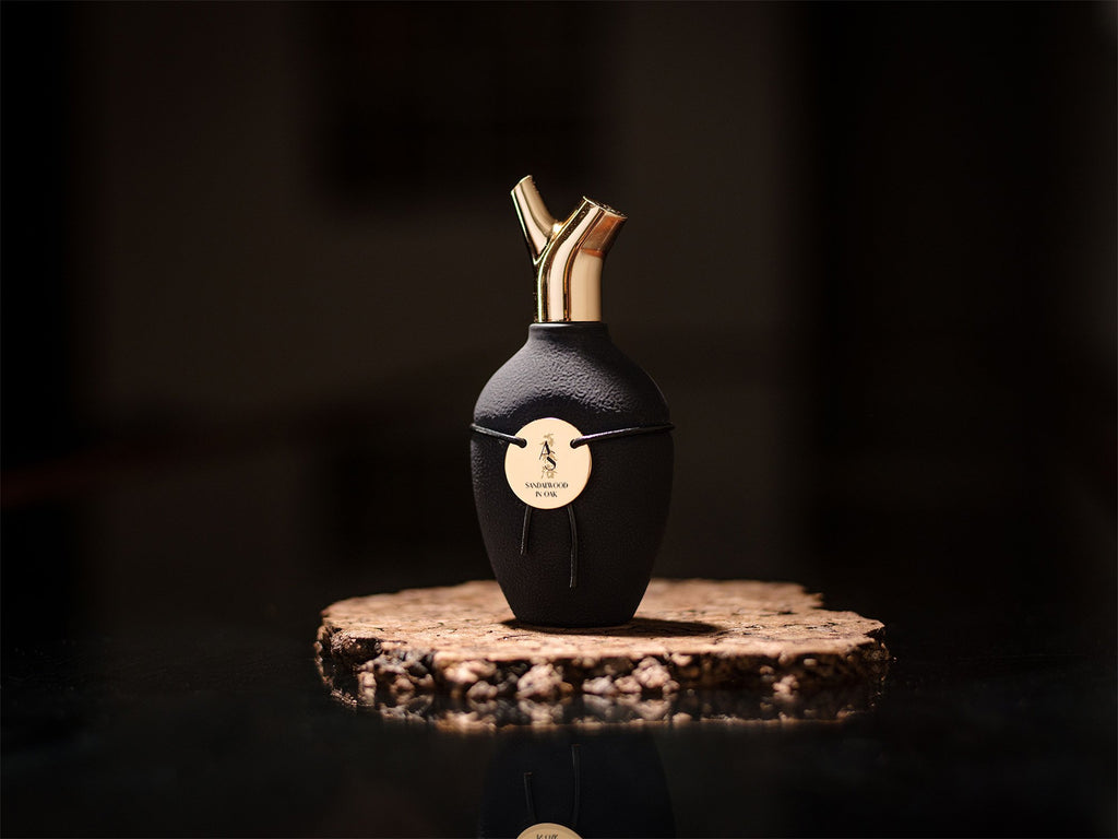 A black bottle with a gold seal and two golden-hued spouts sits on a textured cork base, set against a dark background with hints of Guaiacwood essence. This exquisite piece is named Sandalwood In Oak by Scents of Wood.
