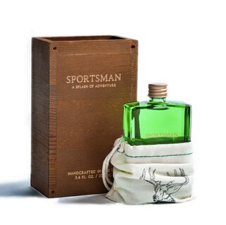 A green glass bottle labeled "Sportsman" by EastWest Bottlers peeks out of a canvas drawstring pouch, adjacent to a wooden box with matching labeling, evoking the rugged scent of the great outdoors.