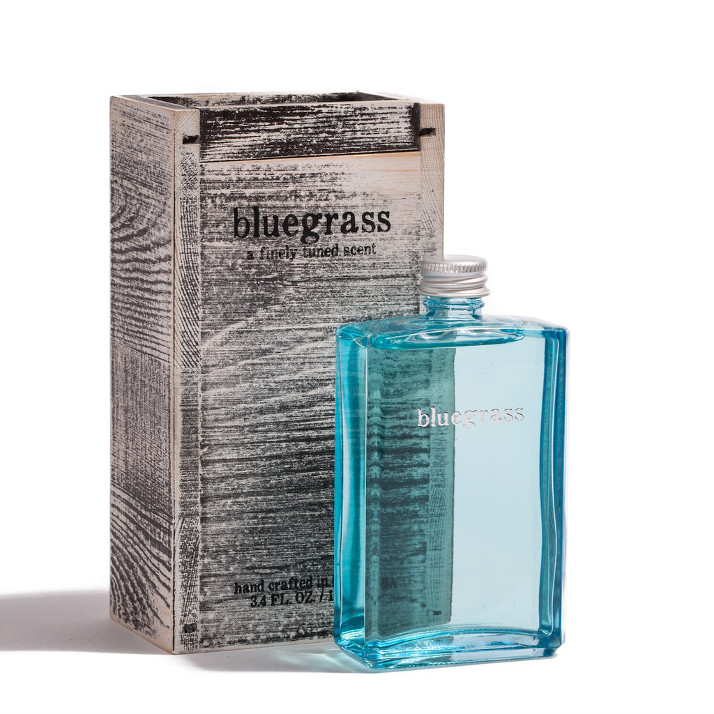 A rectangular, blue-tinted glass bottle labeled "Bluegrass by EastWest Bottlers" stands in front of a rustic, textured box with matching branding.
