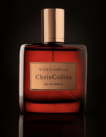 A bottle of Chris Collins Eau de Parfum with a dark red glass body and a gold cap, set against a dark background. The label reads "VIDE COR MEUM Chris Collins," exuding an aura of seduction atmosphere with hints of Armenian style rose.
