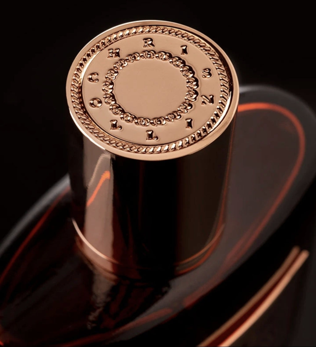 Close-up of a metallic perfume bottle cap featuring the engraved text "Chris Collins" arranged in a circular pattern. The bottle, with its alluring VIDE COR MEUM Eau de Parfum, is dark and reflects light subtly.