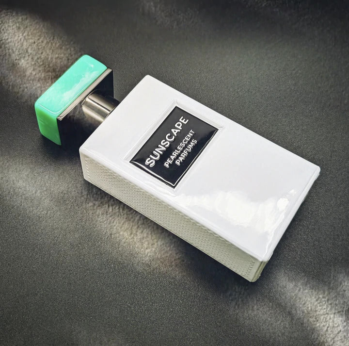 A white perfume bottle with a metallic cap and green accent, labeled "Sunscape" on a black plaque, sits gracefully on a dark gray surface. The scent captures the essence of Texas Sunshine with subtle hints of Solar Citruses. From the collection by Pearlescent Parfums.
