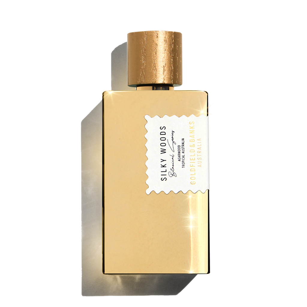 A gold rectangular perfume bottle featuring a matching cylindrical cap is labeled 'Silky Woods' from Goldfield & Banks. The label also highlights 'eau de parfum' and 'hypnotic & sensuous,' making it the epitome of luxury fragrance, enriched with the essence of agarwood.