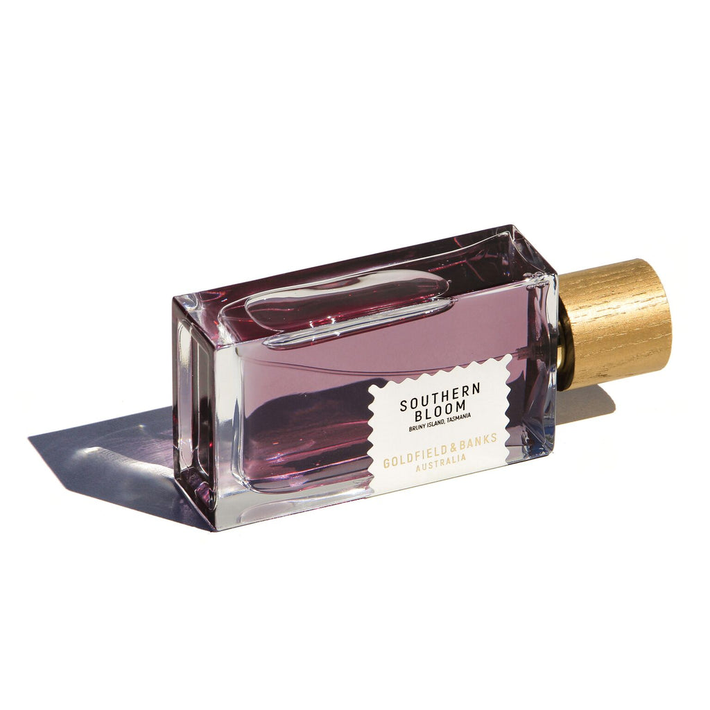 A bottle of Southern Bloom by Goldfield & Banks, showcasing its luxurious fragrance with a purple liquid and a wooden cap, lies elegantly on its side against a white background.