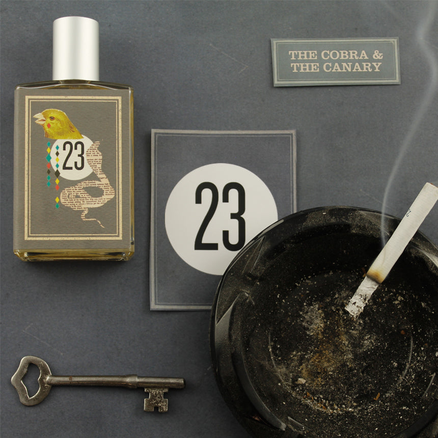 A bottle labeled "23" featuring a bird and snake image, alongside a card marked with "23," an ashtray holding a lit cigarette, a metal key, and an evocative label reading "The Cobra & The Canary" from Imaginary Authors on a gray surface conjure the decadence and desolation of Palm Springs race tracks.