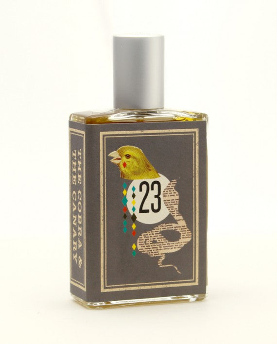 A rectangular perfume bottle from Imaginary Authors, named The Cobra & The Canary, features a gray label adorned with an illustration of a canary and the number "23," evocative of the decadence and desolation of Palm Springs race tracks. A metallic gray cap complements the text on the label that reads, "The Cobra & The Canary.