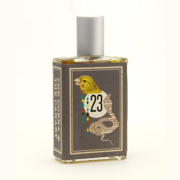 A rectangular perfume bottle from Imaginary Authors, named The Cobra & The Canary, features a gray label adorned with an illustration of a canary and the number "23," evocative of the decadence and desolation of Palm Springs race tracks. A metallic gray cap complements the text on the label that reads, "The Cobra & The Canary.