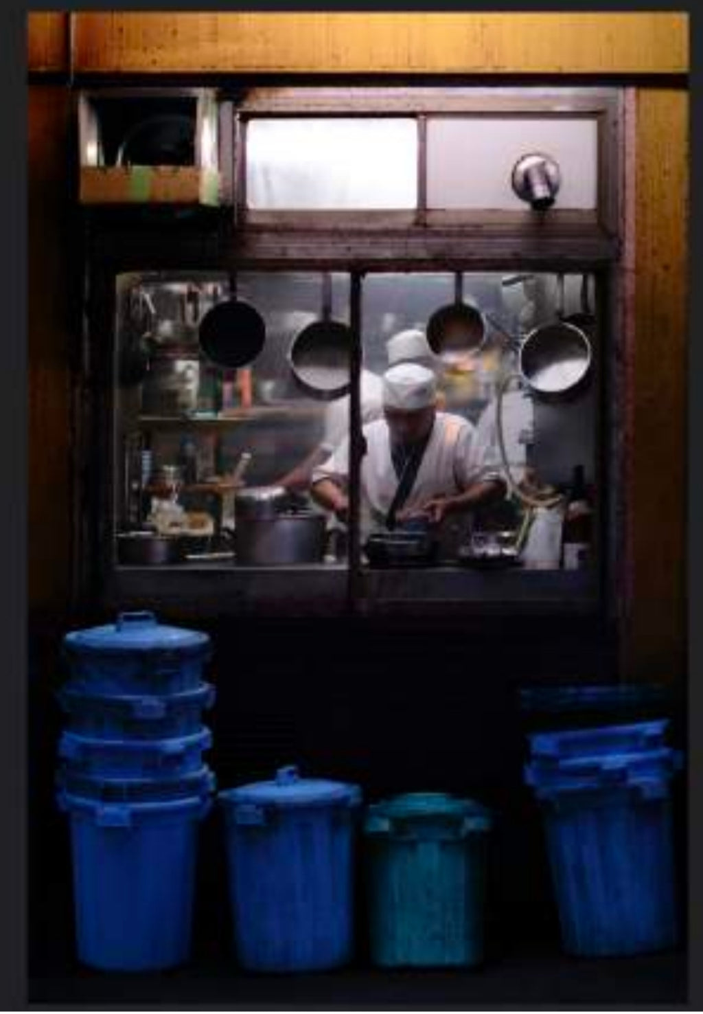 A chef in a white uniform works in a kitchen visible through a window, with multiple cooking pots hanging inside. The air is filled with the zingy, spicy, woody fragrance reminiscent of Tokyo by Gallivant from Gallivant Perfumes. Blue and green trash bins are lined up outside below the window.