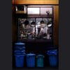 A chef in a white uniform works in a kitchen visible through a window, with multiple cooking pots hanging inside. The air is filled with the zingy, spicy, woody fragrance reminiscent of Tokyo by Gallivant from Gallivant Perfumes. Blue and green trash bins are lined up outside below the window.