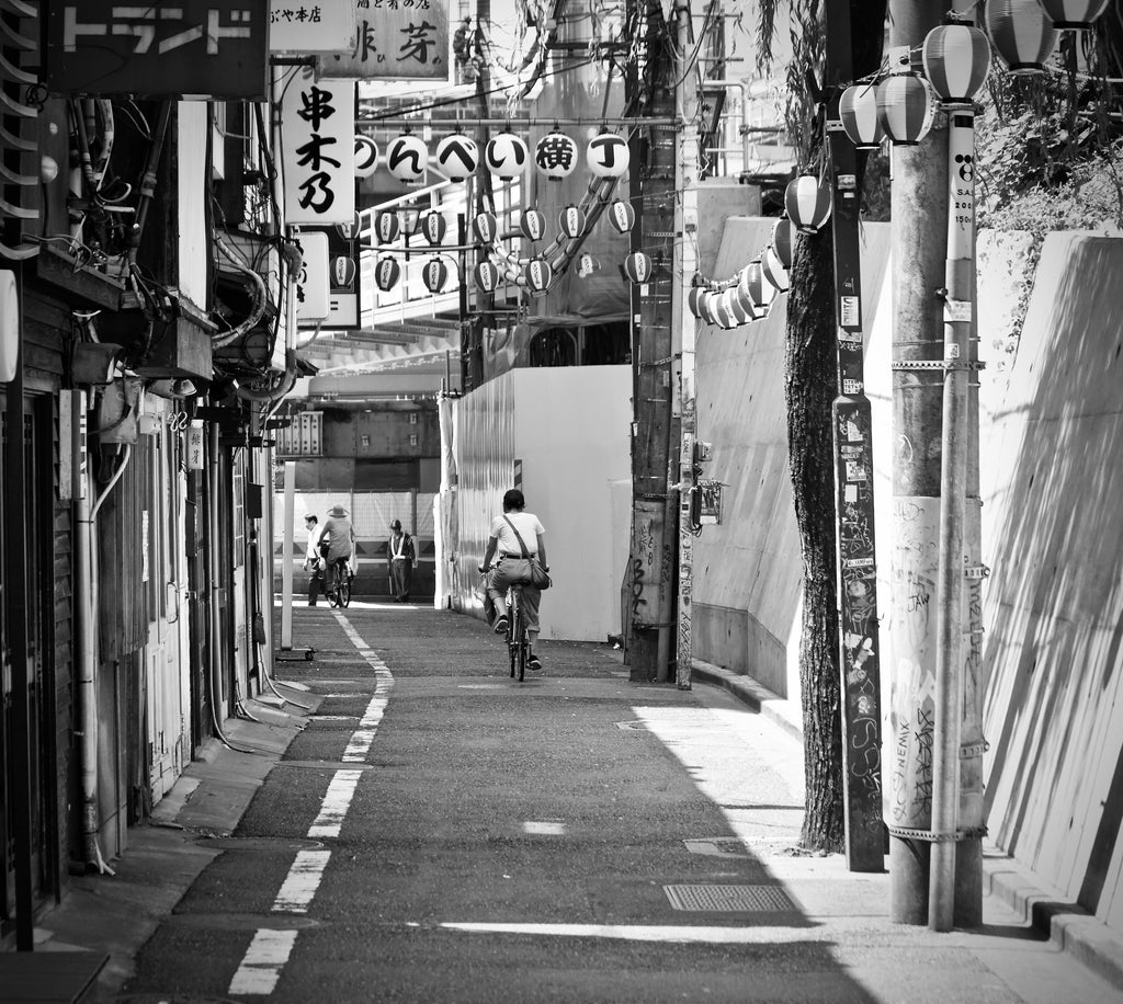 A man rides a bicycle down an alleyway lined with buildings and Japanese signage, while another cyclist and a pedestrian move further ahead. Lanterns hang above the street, their soft glow reminiscent of sandalwood smoky incense wafting through the night, evoking the essence of Tokyo by Gallivant from Gallivant Perfumes. The image is in black and white.