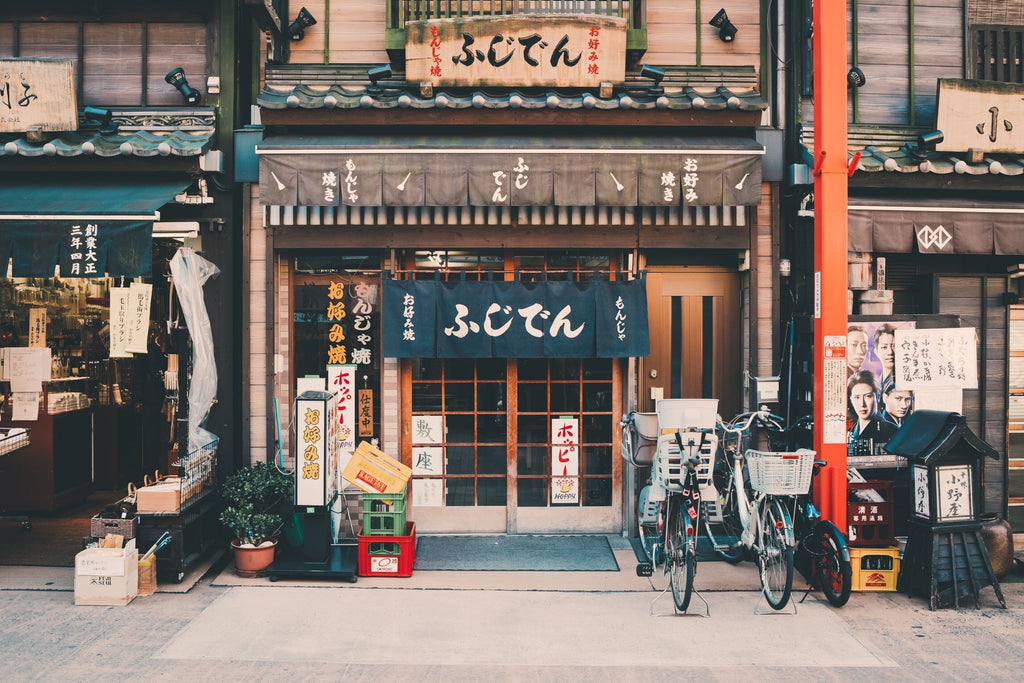 The storefront of a traditional Japanese shop with bicycles parked outside features paper signs and wooden signage. The entrance showcases a fabric curtain adorned with Japanese characters, calling to mind the warm and inviting notes of Tokyo by Gallivant from Gallivant Perfumes, reminiscent of sandalwood smoky incense. Adjacent shops and signs complete the scene.