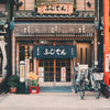 The storefront of a traditional Japanese shop with bicycles parked outside features paper signs and wooden signage. The entrance showcases a fabric curtain adorned with Japanese characters, calling to mind the warm and inviting notes of Tokyo by Gallivant from Gallivant Perfumes, reminiscent of sandalwood smoky incense. Adjacent shops and signs complete the scene.
