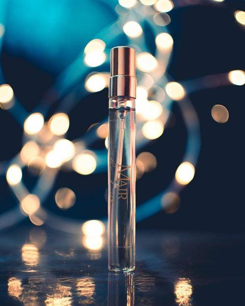 A clear glass bottle of "Remember When" perfume by Mair with a metallic cap stands on a reflective surface, capturing the essence of its citrus floral fragrance. Soft, warm fairy lights in the background create a dreamy, bokeh effect.