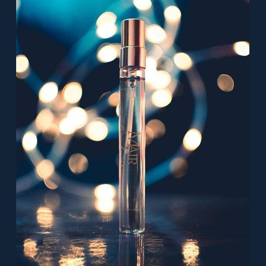 A clear glass bottle of "Remember When" perfume by Mair with a metallic cap stands on a reflective surface, capturing the essence of its citrus floral fragrance. Soft, warm fairy lights in the background create a dreamy, bokeh effect.