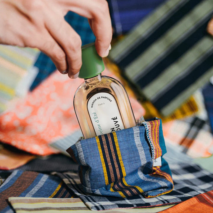 A hand holds a bottle of Verveine d'Eté by Le Jardin Retrouvé with a green cap, partially inserted into a striped cloth pocket, with colorful fabric patterns in the background.