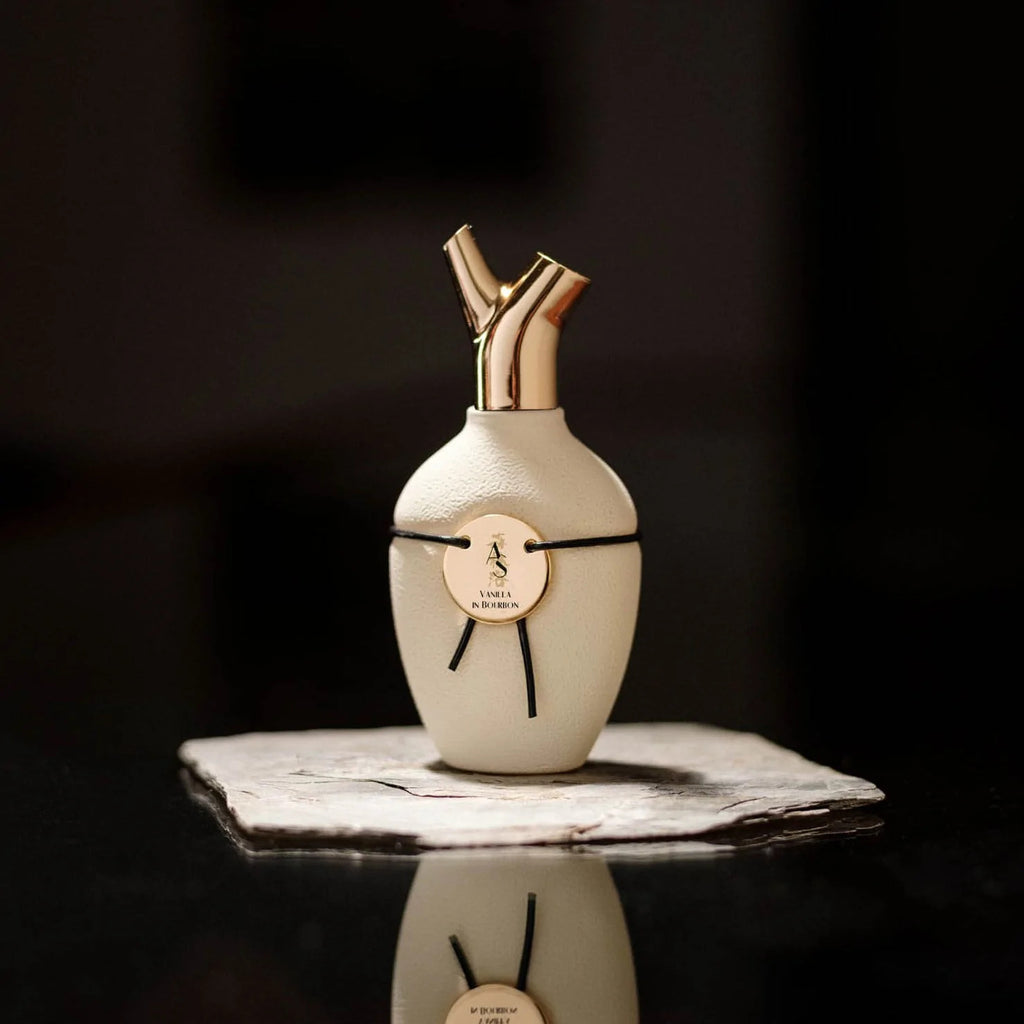 A white, textured bottle of Vanilla in Bourbon Eau de Toilette by Scents of Wood with gold dual spouts sits on a stone slab, adorned with a black ribbon and round gold emblem. The scent harmonizes notes of vanilla with hints of myrrh resoid, creating an exquisite fragrance experience.