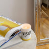Lit candle in white jar with yellow band, VISION label on front