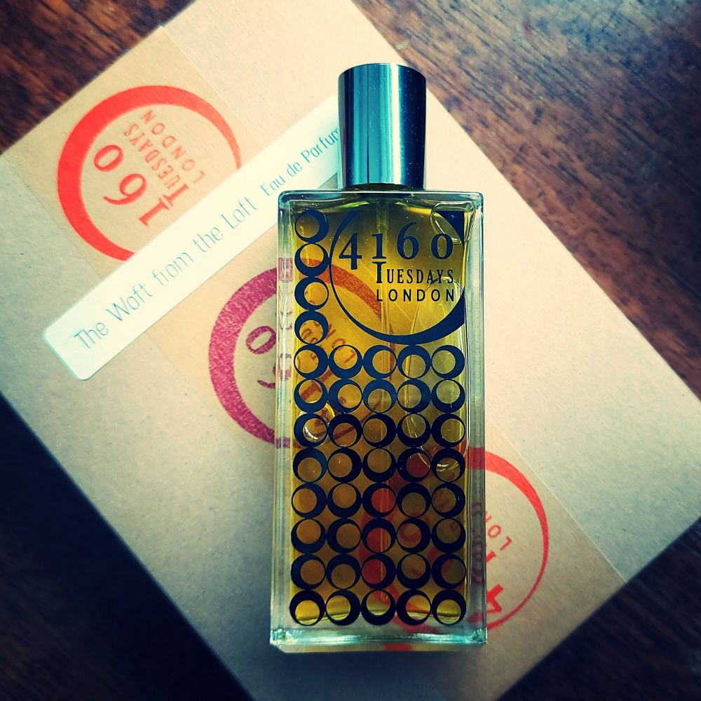 A perfume bottle labeled "4160Tuesdays" is placed on top of a brown package with red stamps and a white label reading "The Waft from the Loft Limited Edition Eau de Parfum.