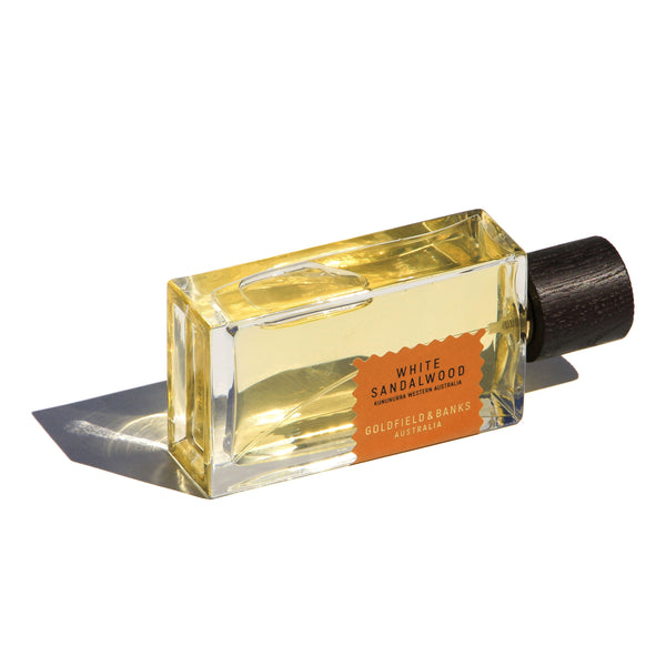 A rectangular bottle of Goldfield & Banks White Sandalwood perfume, filled with yellow liquid, is adorned with a label that teases notes of Turkish rose. The sleek black cap casts a shadow on the surface, embodying an aura of elegance infused with amber.