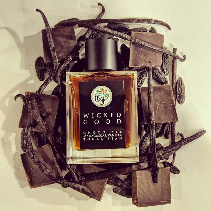 A bottle of "Wicked Good" fragrance by Gallagher Fragrances, surrounded by pieces of milk chocolate and vanilla beans, on a white surface.