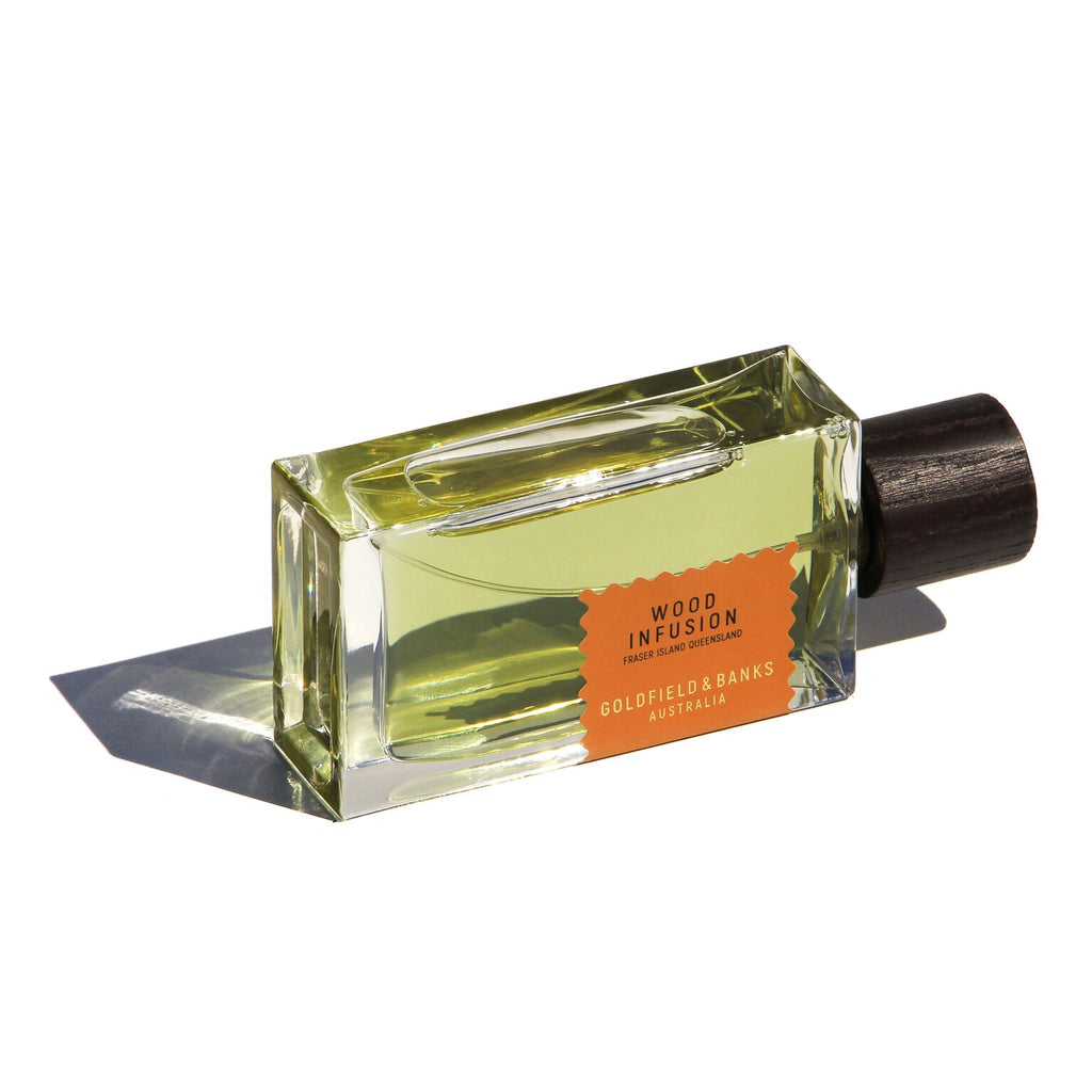 A rectangular glass perfume bottle filled with yellow liquid, labeled "Wood Infusion" and "Goldfield & Banks" on an orange label with a dark wood-textured cap, exudes the rich scent of Australian Exotic Woods.