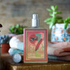 A bottle of Yesterday Haze by Imaginary Authors, with a seductive scent and decorative label, sits on a wooden surface amidst small potted plants and stacked towels.