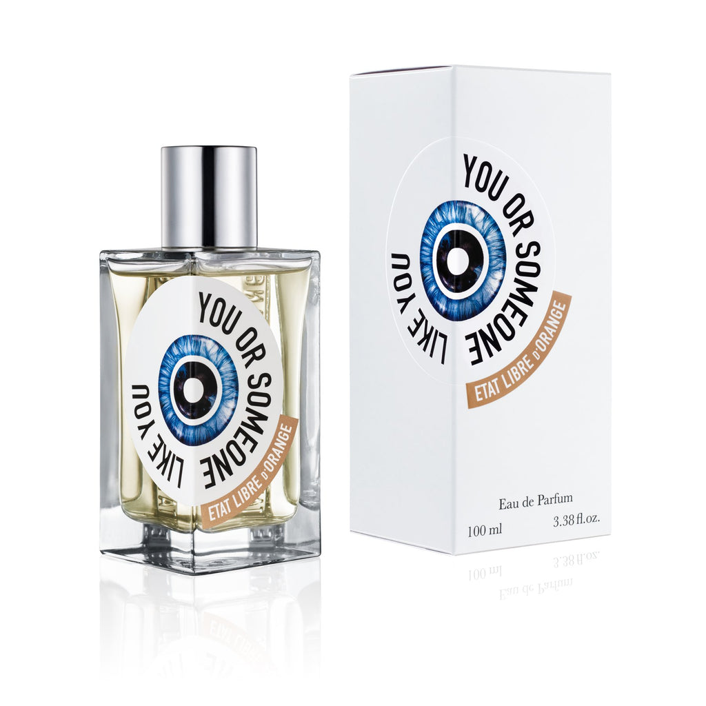 A bottle of "You or Someone Like You" perfume by État Libre d'Orange is placed next to its packaging. The box, with its eye graphic and orange label, hints at the fresh, floral fragrance inside. The clear bottle with a silver cap embodies the sophisticated touch inspired by Chandler Burr.