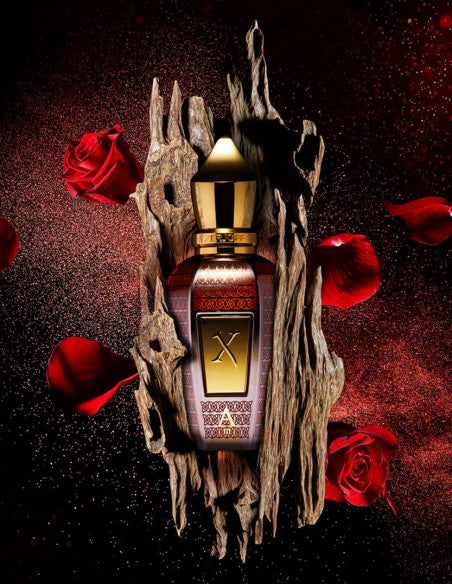 A decorative bottle of Alexandria III by Xerjoff with a gold cap is surrounded by pieces of Laotian Oud and floating red Bulgarian Rose petals against a dark, textured background.