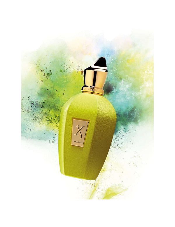 A lime-green Xerjoff Amabile perfume bottle with a black cap and gold label, suspended with a colorful burst of yellow, green, and blue hues in the background, exudes the essence of avant-garde perfumes infused with fresh fruits.