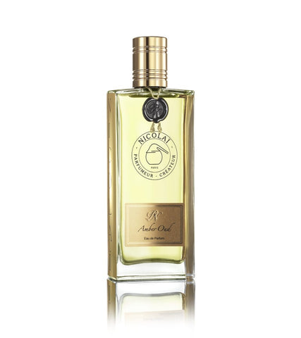 A rectangular bottle of Nicolaï Amber Oud Eau de Parfum with a gold cap and label, displaying a logo and text. The bottle contains a yellow-tinted liquid reminiscent of amber and is set against a white background.