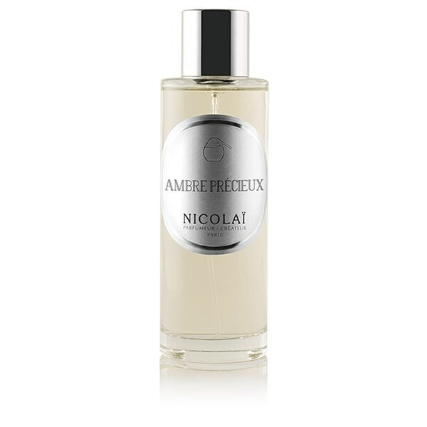 A clear bottle of Nicolaï Ambre Precieux Room Spray with a metallic label and silver cap, containing a yellowish liquid infused with hints of amber.