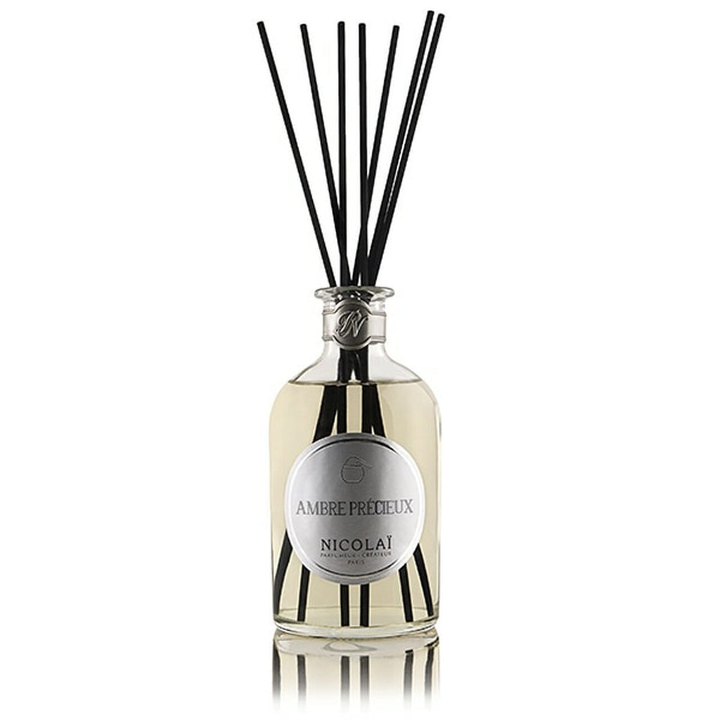 A bottle of Ambre Precieux Reed Diffuser by Nicolaï, a luxurious Amber reed diffuser with a sandalwood vanilla fragrance, complete with several black reeds inserted into the 250ml bottle of clear liquid lasting up to 3 months.