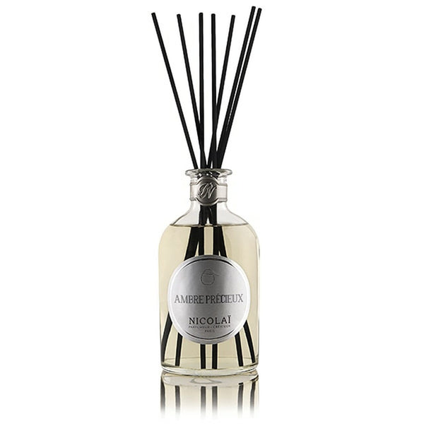 A bottle of Ambre Precieux Reed Diffuser by Nicolaï, a luxurious Amber reed diffuser with a sandalwood vanilla fragrance, complete with several black reeds inserted into the 250ml bottle of clear liquid lasting up to 3 months.