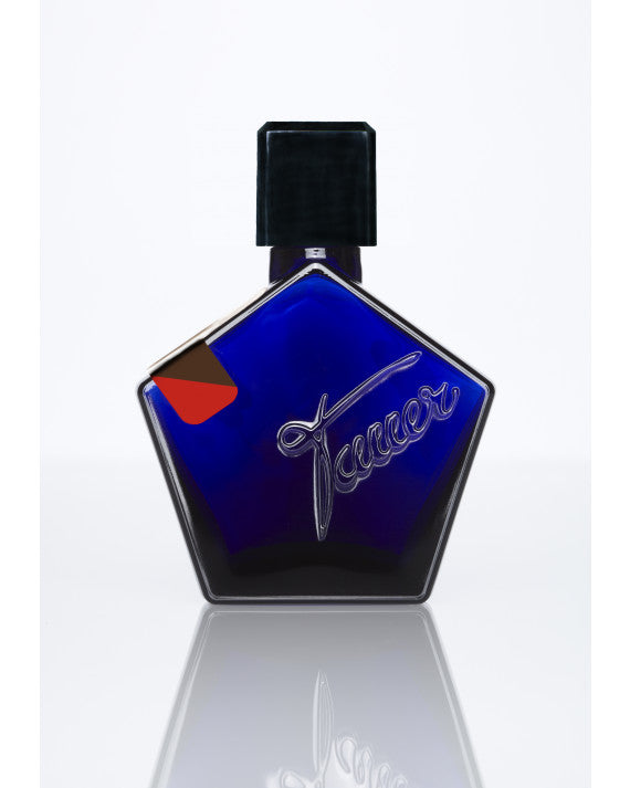 A blue pentagon-shaped perfume bottle with a black cap and cursive script detail, reflecting on a white surface, encapsulating the essence of Au Coeur Du Desert by Tauer Perfumes.